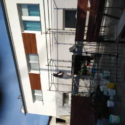 Pracownik na rusztowaniu maluje białą elewację budynku z drewnopodobnymi panelami, widoczne liczne puste i częściowo puste wiadra po farbie, oraz stojące na ziemi deski.