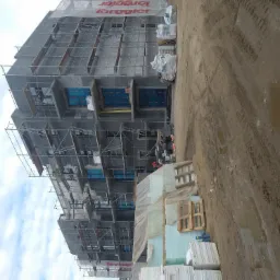 Budynek w trakcie budowy z widocznymi rusztowaniami, częściowo ocieplony, z materiałami budowlanymi składowanymi obok oraz błotnistym terenem.