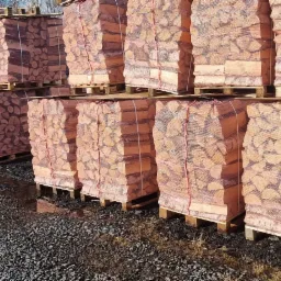 Ułożone na paletach worki z pociętym drewnem kominkowym, zabezpieczone siatką z czerwonym sznurkiem, na żwirowym podłożu.