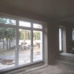 Wnętrze w trakcie prac wykończeniowych: ściany po nałożeniu szarego tynku, widoczne duże okno z widokiem na ogród, w tle schody i dodatkowe okno.