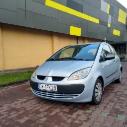 Jasnoniebieski Mitsubishi Colt parkuje na kostce brukowej przed budynkiem z żółto-szarą fasadą w geometryczne wzory. Tablica rejestracyjna z widocznym adresem strony internetowej.