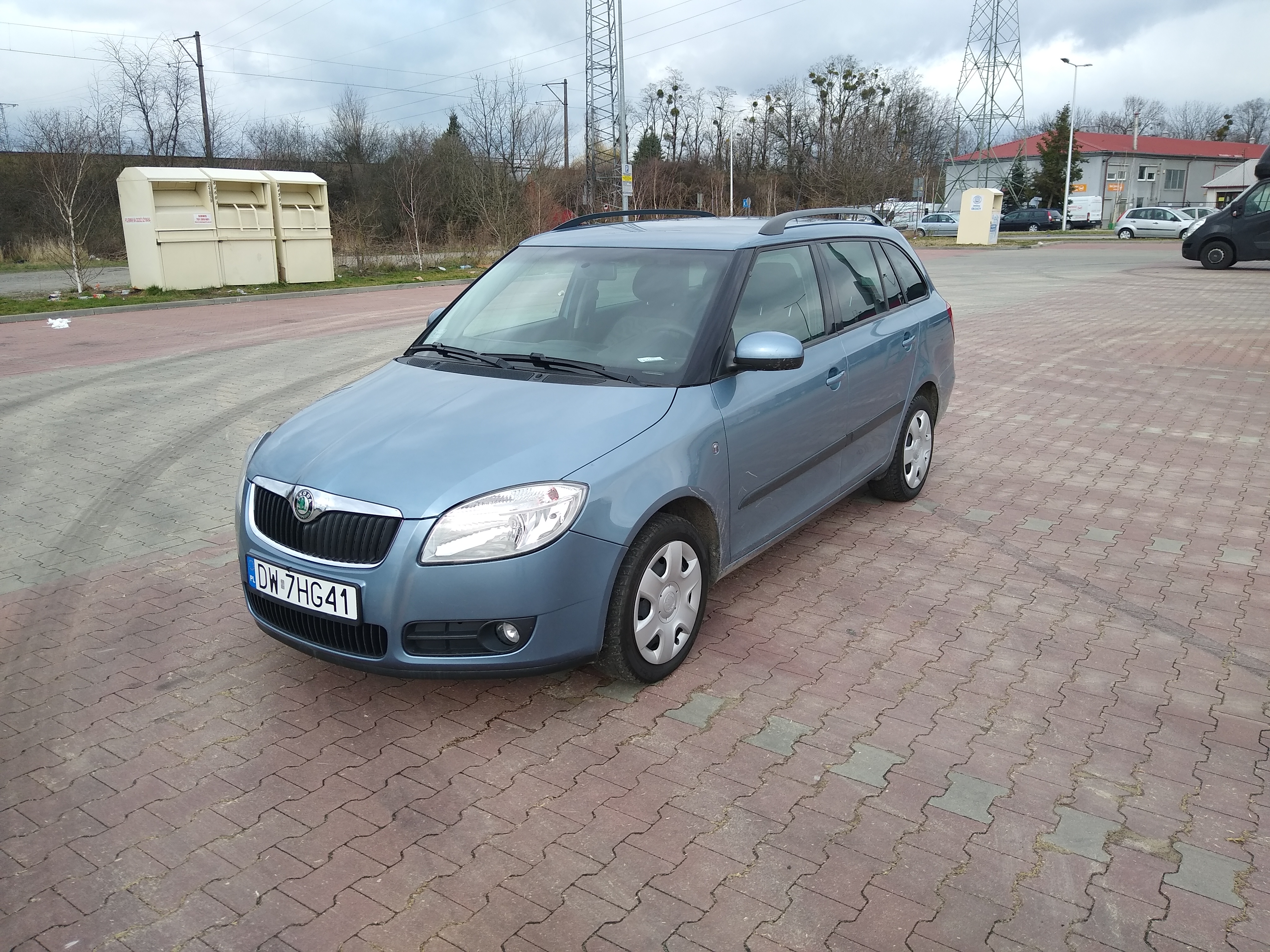 Błękitna Skoda Fabia kombi zaparkowana na brukowanym placu, widoczne kontenery na odzież i słupy wysokiego napięcia w tle.