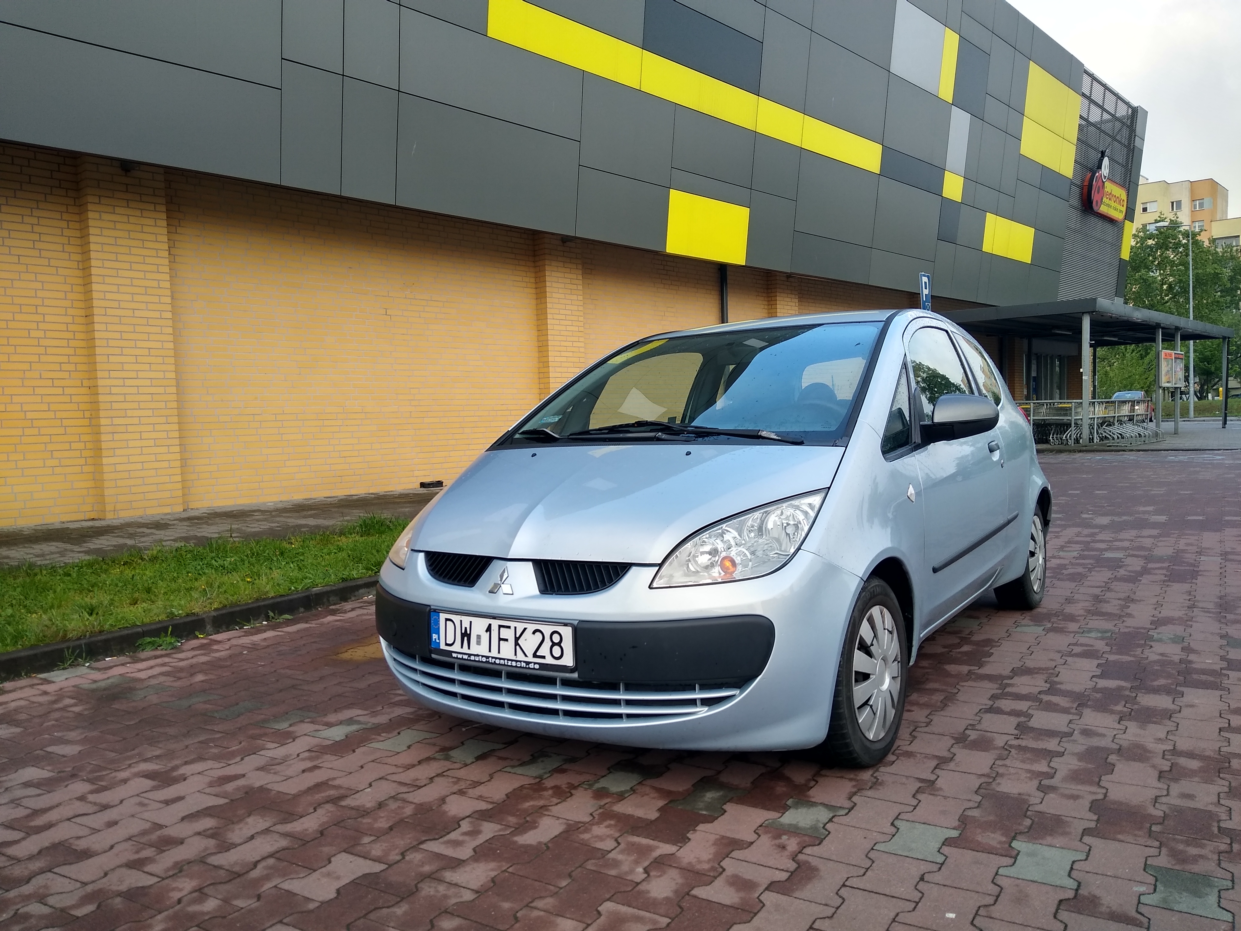 Jasnoniebieski Mitsubishi Colt parkuje na kostce brukowej przed budynkiem z żółto-szarą fasadą w geometryczne wzory. Tablica rejestracyjna z widocznym adresem strony internetowej.