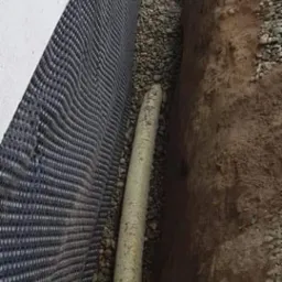 Rów wykopany w ziemi, wyłożony czarną folią z otworami, z rurą drenażową na dnie, zasypany drobnymi kamieniami.