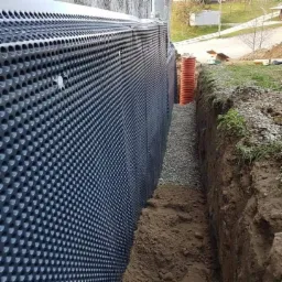 Czarna folia kubełkowa z otworami izolująca fundament budynku, zasypana żwirem, z widoczną rurą drenarską w wykopie.