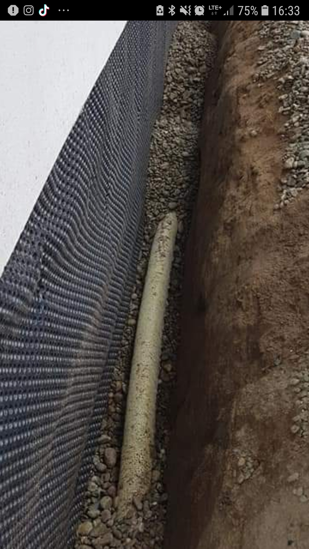 Rów wykopany w ziemi, wyłożony czarną folią z otworami, z rurą drenażową na dnie, zasypany drobnymi kamieniami.