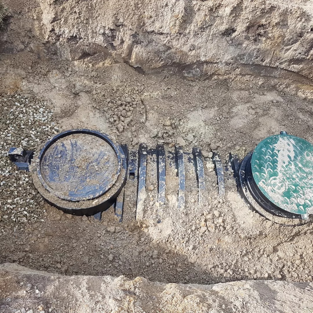 Studzienka kanalizacyjna z pokrywą w wykopie, widoczne elementy systemu drenażowego i żwir.