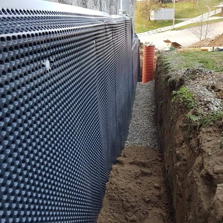 Czarna folia kubełkowa z otworami izolująca fundament budynku, zasypana żwirem, z widoczną rurą drenarską w wykopie.