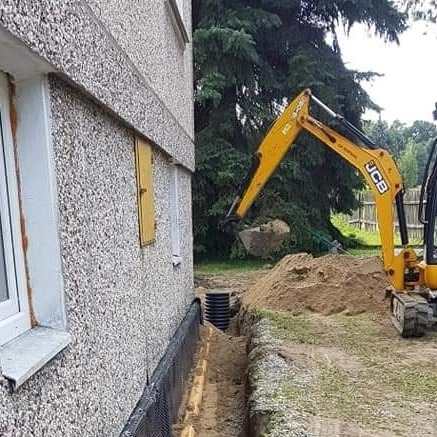 Żółta koparka JCB zasypuje wykop pod fundamentem budynku, widoczne rury drenażowe i izolacja pionowa ściany fundamentowej.