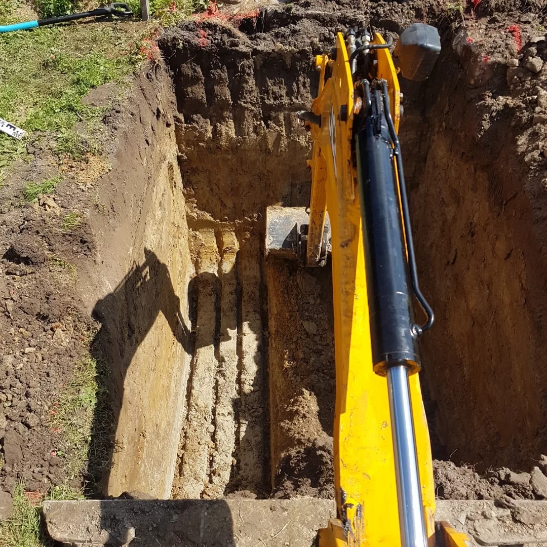 Żółta koparka w trakcie wykonywania wykopu pod instalacje, widoczne równe ściany wykopu i cień ramienia koparki.