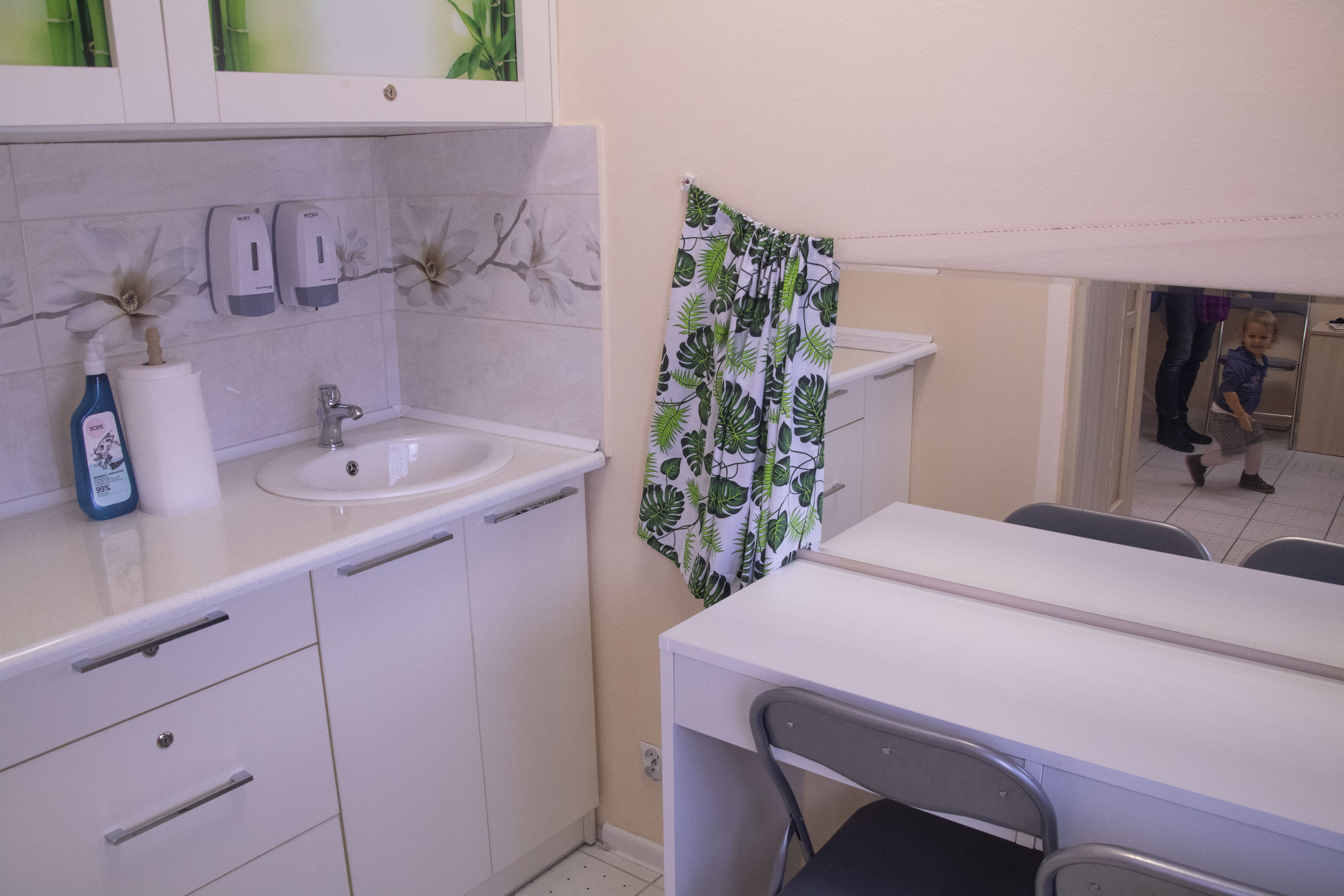 Wnętrze gabinetu z umywalką, szafkami w kolorze złamanej bieli, dozownikami mydła i ręczników, oraz lustrem odbijającym bawiące się dziecko.