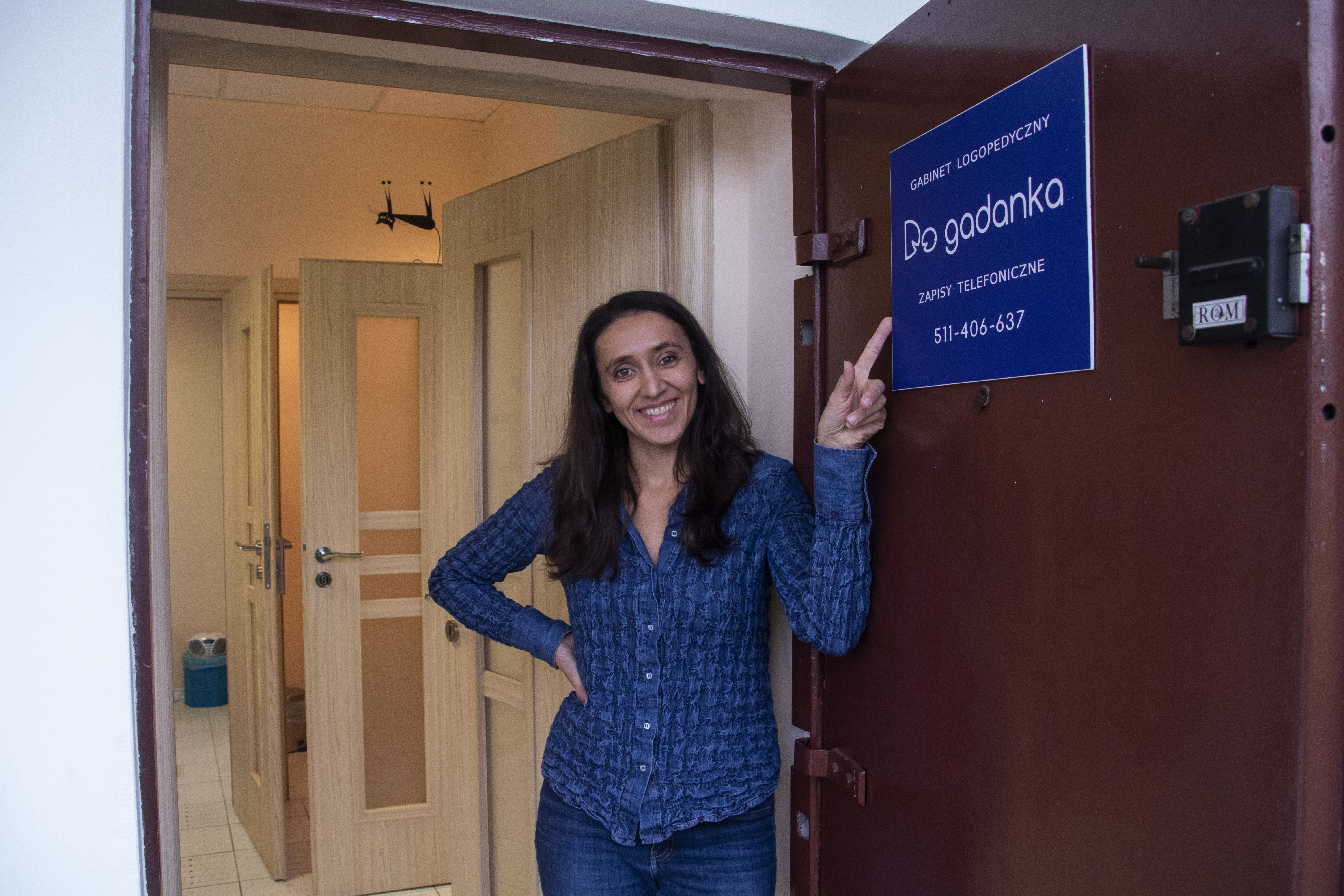 Uśmiechnięta kobieta wskazuje na niebieską tabliczkę z napisem 'Gabinet logopedyczny Do gadanka' i numerem telefonu na ciemnoczerwonych drzwiach gabinetu, widoczne otwarte drzwi do wnętrza.