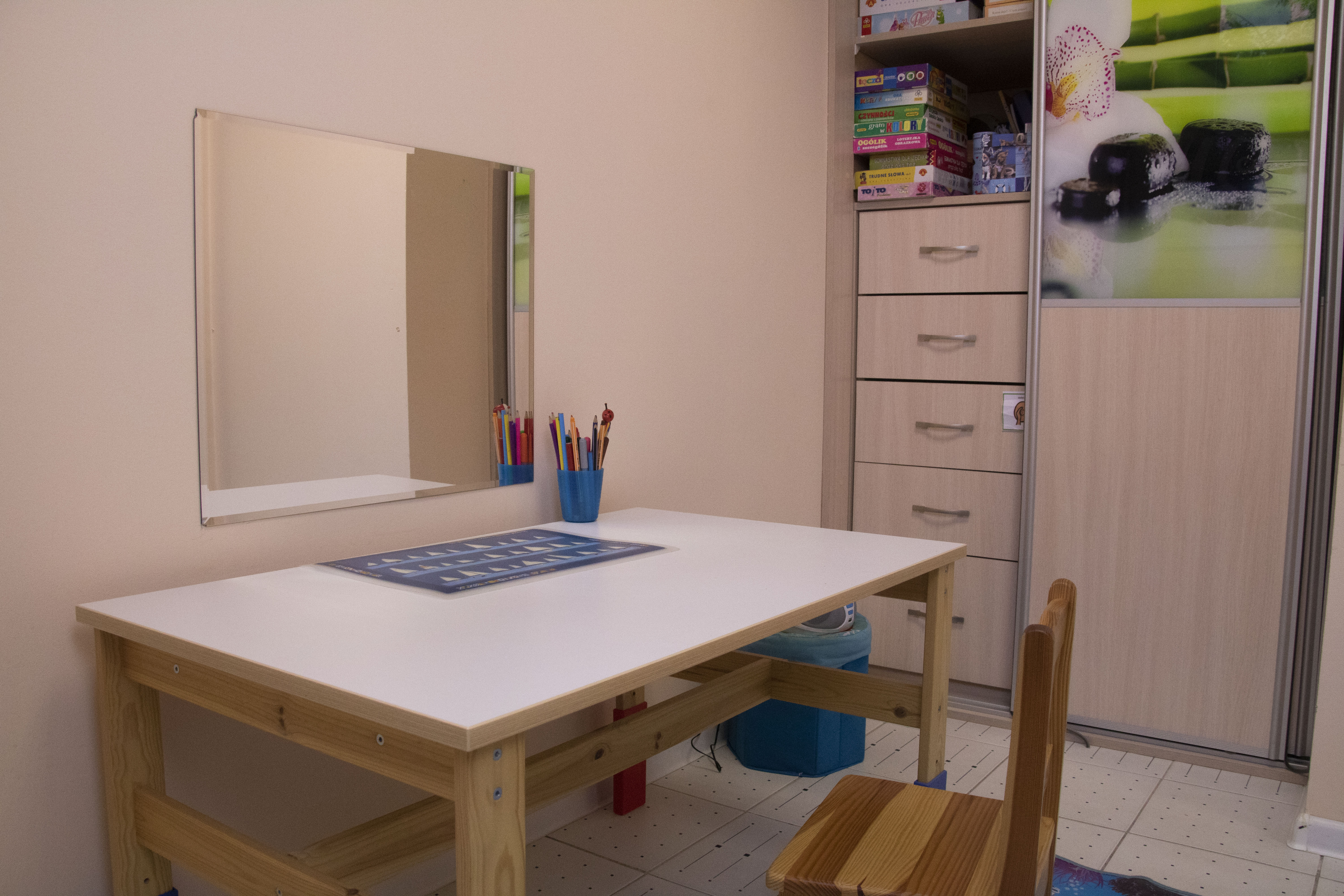 Gabinet terapeutyczny z biurkiem, krzesłem i lustrem, na biurku kolorowe kredki i plansza edukacyjna, w tle szafa z grami i dekoracją zen.