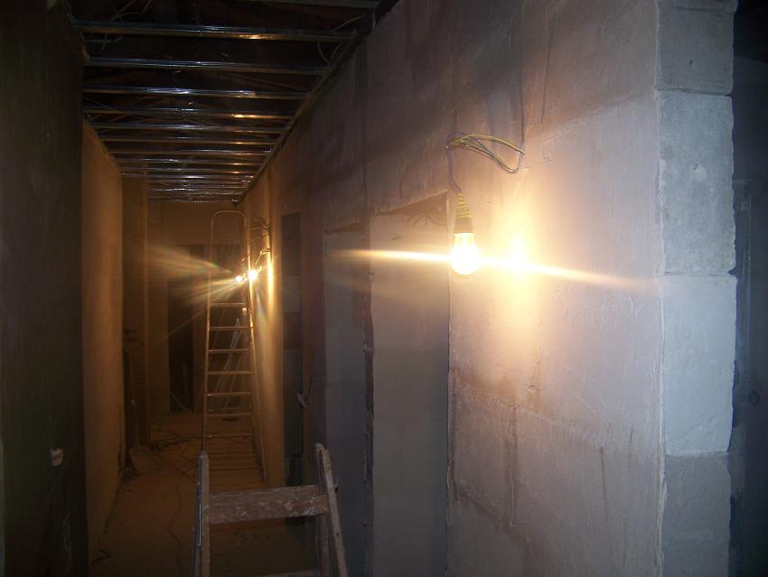 Wnętrze w trakcie budowy ścian działowych z widoczną konstrukcją sufitu podwieszanego, oświetlone wiszącą żarówką, z drabina w tle.
