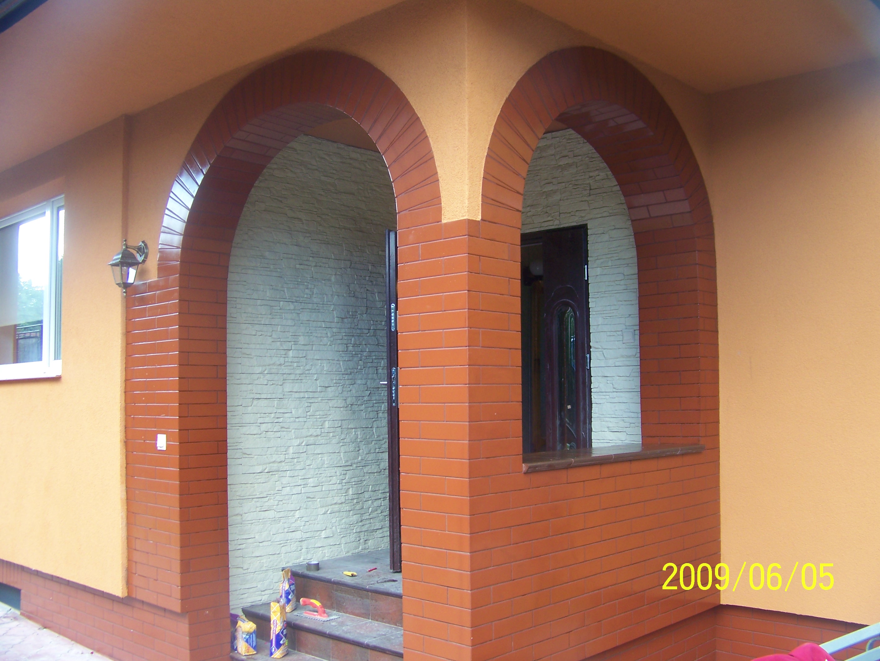 Wykończone wejście do budynku z dwoma łukowymi przejściami obłożonymi czerwoną cegłą, ściany pomalowane na pomarańczowo, wewnątrz widoczna ściana wyłożona dekoracyjnym kamieniem, na schodach leżą...