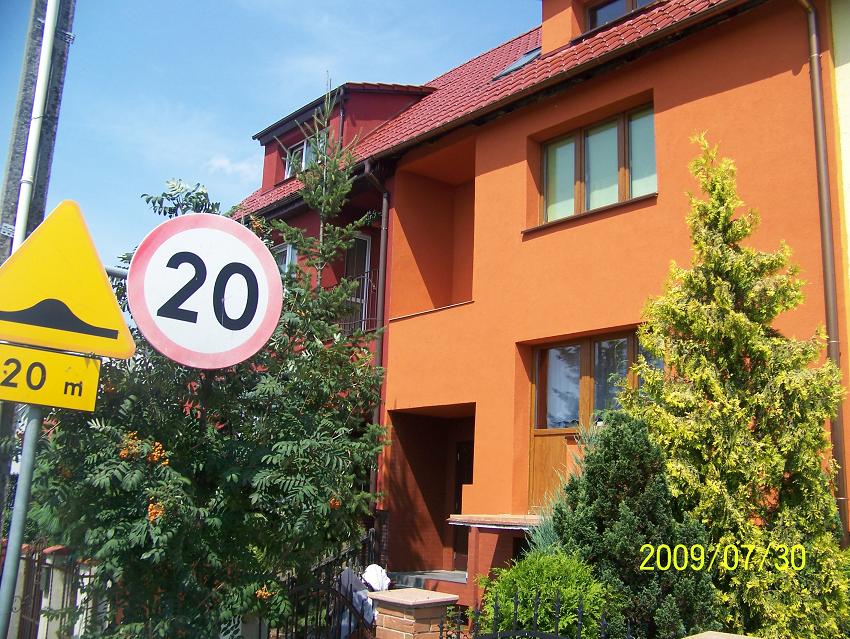 Pomarańczowy dom jednorodzinny z czerwoną dachówką, widoczny znak drogowy ograniczenia prędkości do 20 km/h oraz znak ostrzegawczy o progu zwalniającym, zieleń wokół budynku, data wykonania zdjęcia...