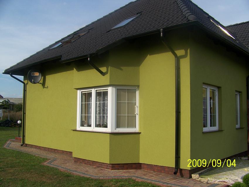 Dom jednorodzinny po dociepleniu elewacji, pomalowany na oliwkowy kolor, z białymi oknami i ciemnym dachem z lukarnami. Widoczna antena satelitarna i fragment chodnika z kostki.