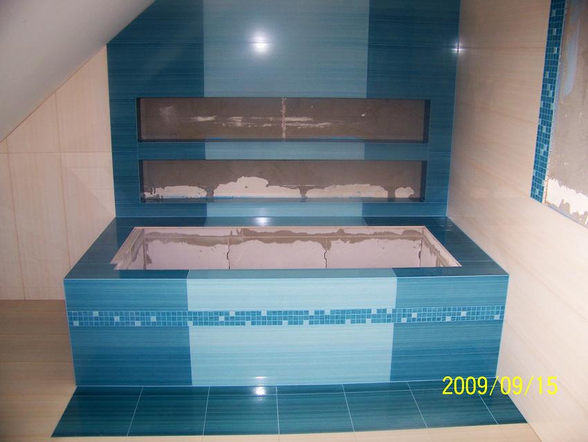 Niedokończona zabudowa wnęki wannowej z niebieskich płytek w różnych odcieniach, widoczne dwie puste wnęki na ścianie i fragment daty na dole zdjęcia.