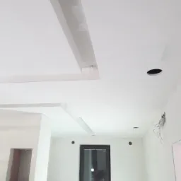 Wnętrze w trakcie prac wykończeniowych: białe ściany, sufit z dekoracyjnymi elementami gipsowymi, okno z czarną ramą, widoczne przewody elektryczne i otwory na oświetlenie, drabina oparta o ścianę.