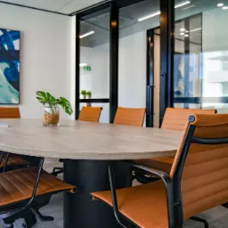 Nowoczesna sala konferencyjna z owalnym stołem z blatem w kolorze jasnego drewna, obitymi brązową skórą krzesłami na czarnych nogach, obrazem w odcieniach niebieskiego i zielonego na ścianie...