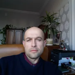 Portret mężczyzny w swetrze na tle biura z roślinami, szafką i tapetą w kwiaty.