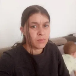 Kobieta o powściągliwym wyrazie twarzy, w ciemnej bluzce, siedząca na kanapie. W tle widoczna głowa dziecka. Zdjęcie z bliska.