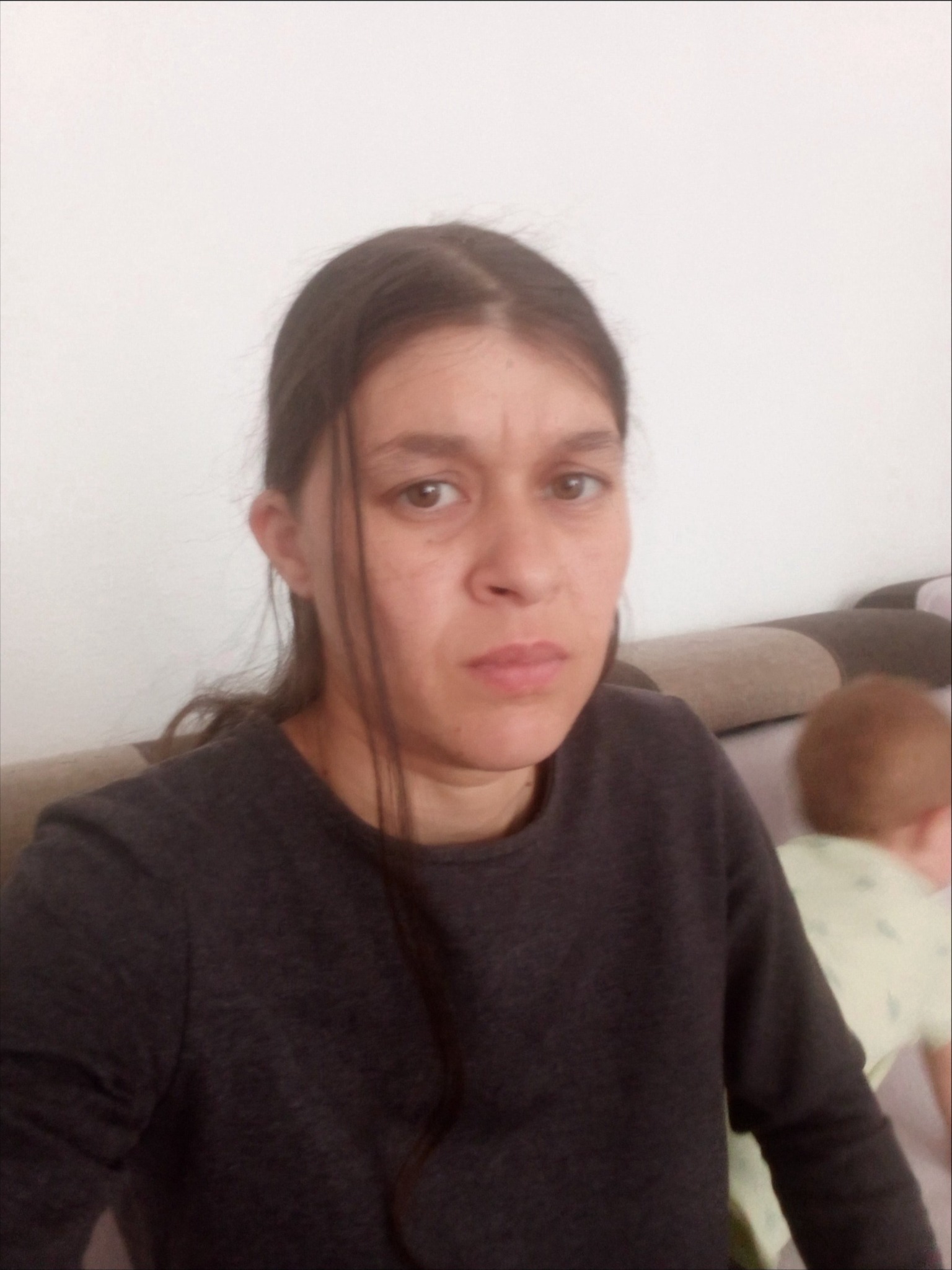 Kobieta o powściągliwym wyrazie twarzy, w ciemnej bluzce, siedząca na kanapie. W tle widoczna głowa dziecka. Zdjęcie z bliska.