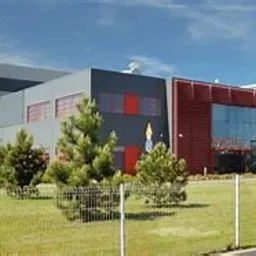 Szeroki, panoramiczny widok na nowoczesny budynek biurowy z szarymi i czerwonymi elementami fasady, otoczony zielonym trawnikiem, drzewami iglastymi i metalowym ogrodzeniem na pierwszym planie,...