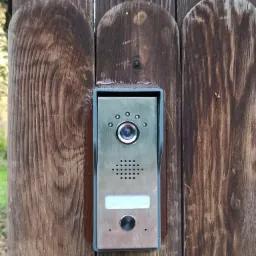 Metalowy panel wideodomofonu z kamerą, głośnikiem i przyciskiem dzwonka zamontowany na brązowym, drewnianym ogrodzeniu z pionowych desek z zaokrąglonymi końcami.