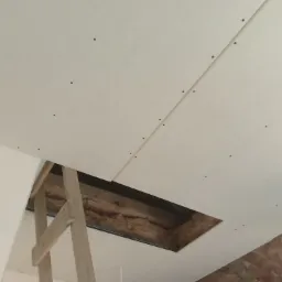 Montaż płyt gipsowo-kartonowych na suficie, widoczne wkręty i otwór z ociepleniem, drewniana konstrukcja wspornikowa.