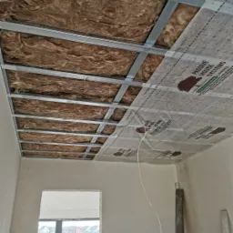 Montaż izolacji z wełny mineralnej i folii paroizolacyjnej z nadrukiem Passive House Systems na metalowym ruszcie sufitu podwieszanego, widoczne wystające kable instalacji elektrycznej.