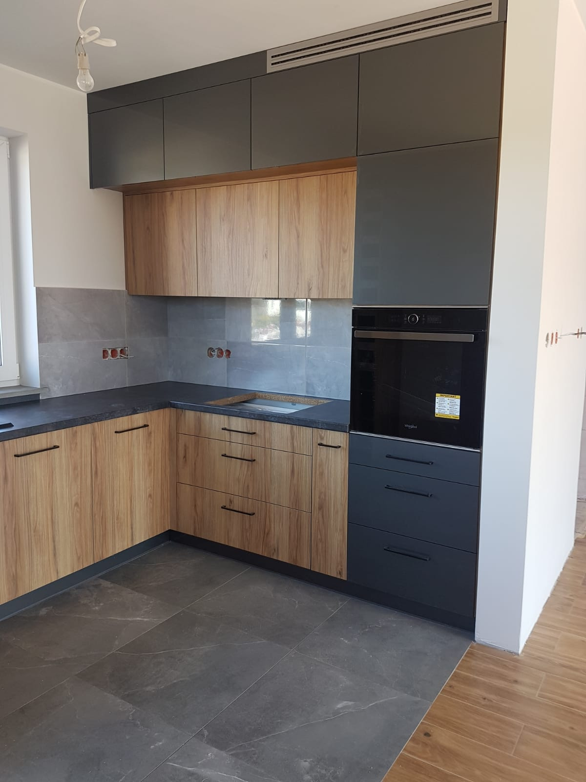 Nowoczesna kuchnia z drewnianymi frontami szafek dolnych i szarymi górnymi, z wbudowanym piekarnikiem i szarymi płytkami na ścianie, widok na fragment podłogi z szarych płytek.