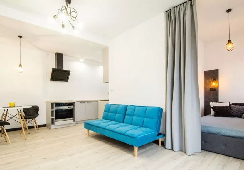 Wnętrze małego mieszkania z aneksem kuchennym, sypialnią oddzieloną zasłoną i turkusową sofą na drewnianych nóżkach, oświetlone lampami w stylu industrialnym.