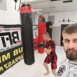 Selfie mężczyzny w koszulce z logo klubu sportów walki na tle sali treningowej z workami bokserskimi i trenującym zawodnikiem.