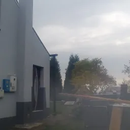 Świeżo otynkowana elewacja domu jednorodzinnego w szary odcień, z widocznymi blokami styropianu i deskami na trawniku, pochmurne niebo w tle.