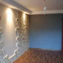 Pokój z dekoracyjną ścianą z kamienia łupanego, podświetloną reflektorem, z drewnianą podłogą i szarą ścianą z drewnianymi drzwiami.