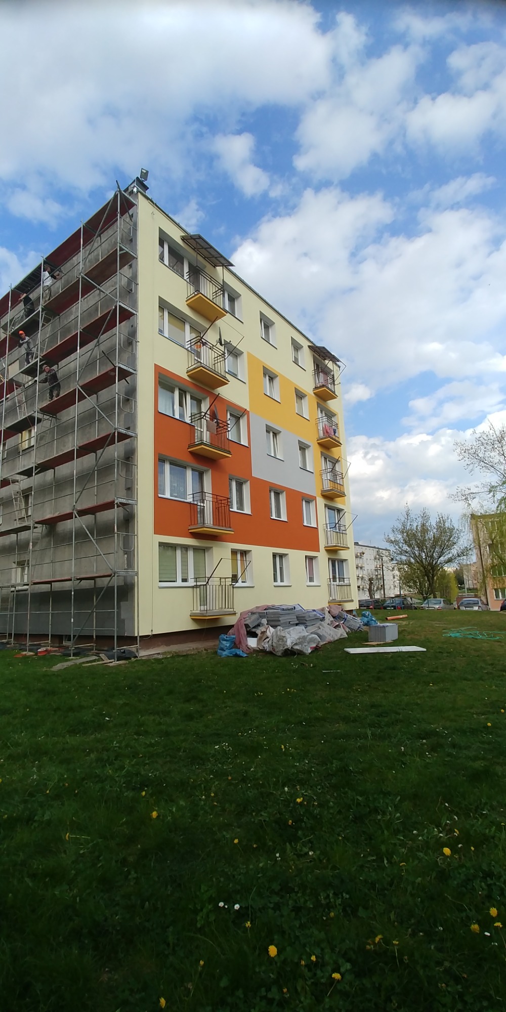 Wielorodzinny budynek w trakcie odnawiania elewacji, widoczne rusztowania, fragmenty ocieplonej i pomalowanej ściany w odcieniach żółci, pomarańczy i szarości, na trawniku leżą materiały budowlane.