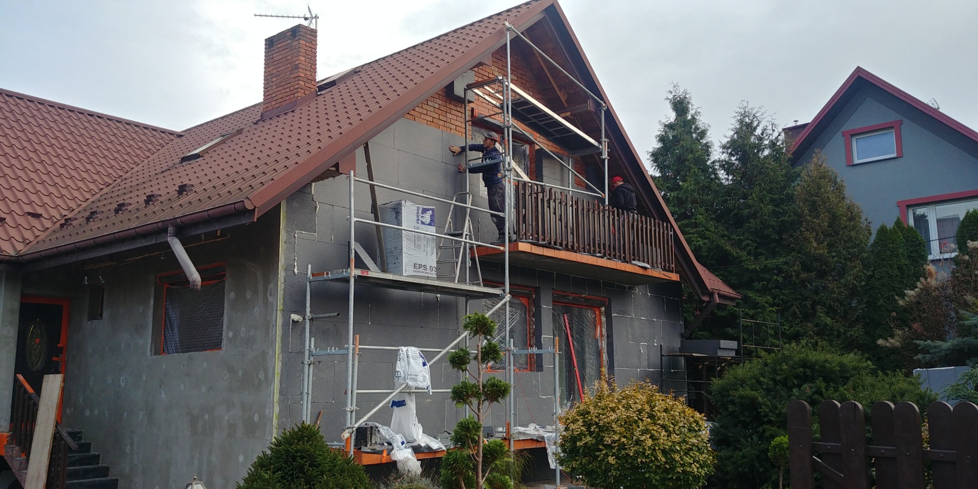 Dom w trakcie ocieplania z użyciem styropianu EPS 03, widoczne rusztowania, pracownicy montujący izolację oraz fragment otynkowanej ściany.