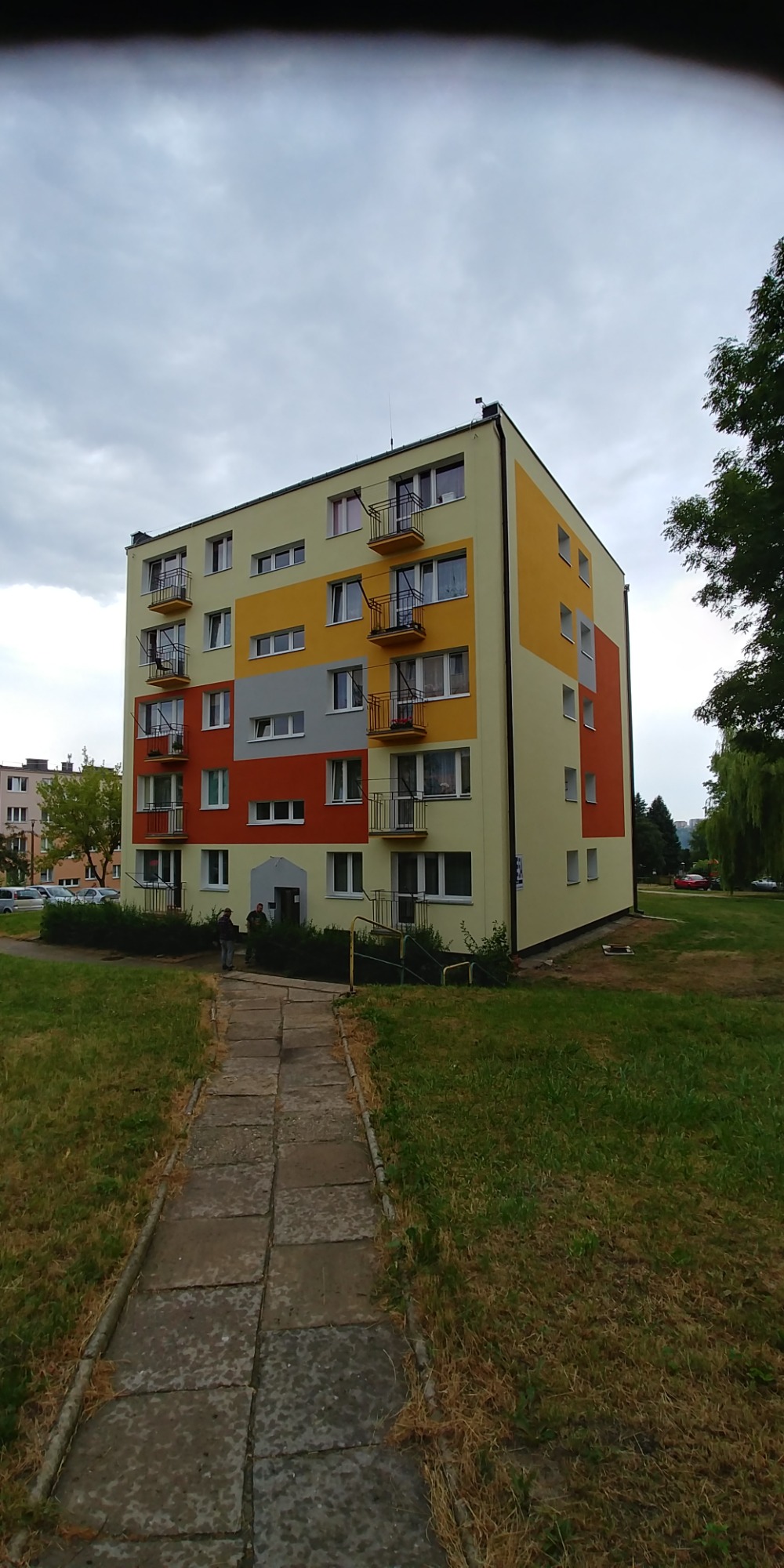 Wielobarwny blok mieszkalny po renowacji elewacji, z balkonami i chodnikiem prowadzącym do wejścia, widok z poziomu trawnika w pochmurny dzień.