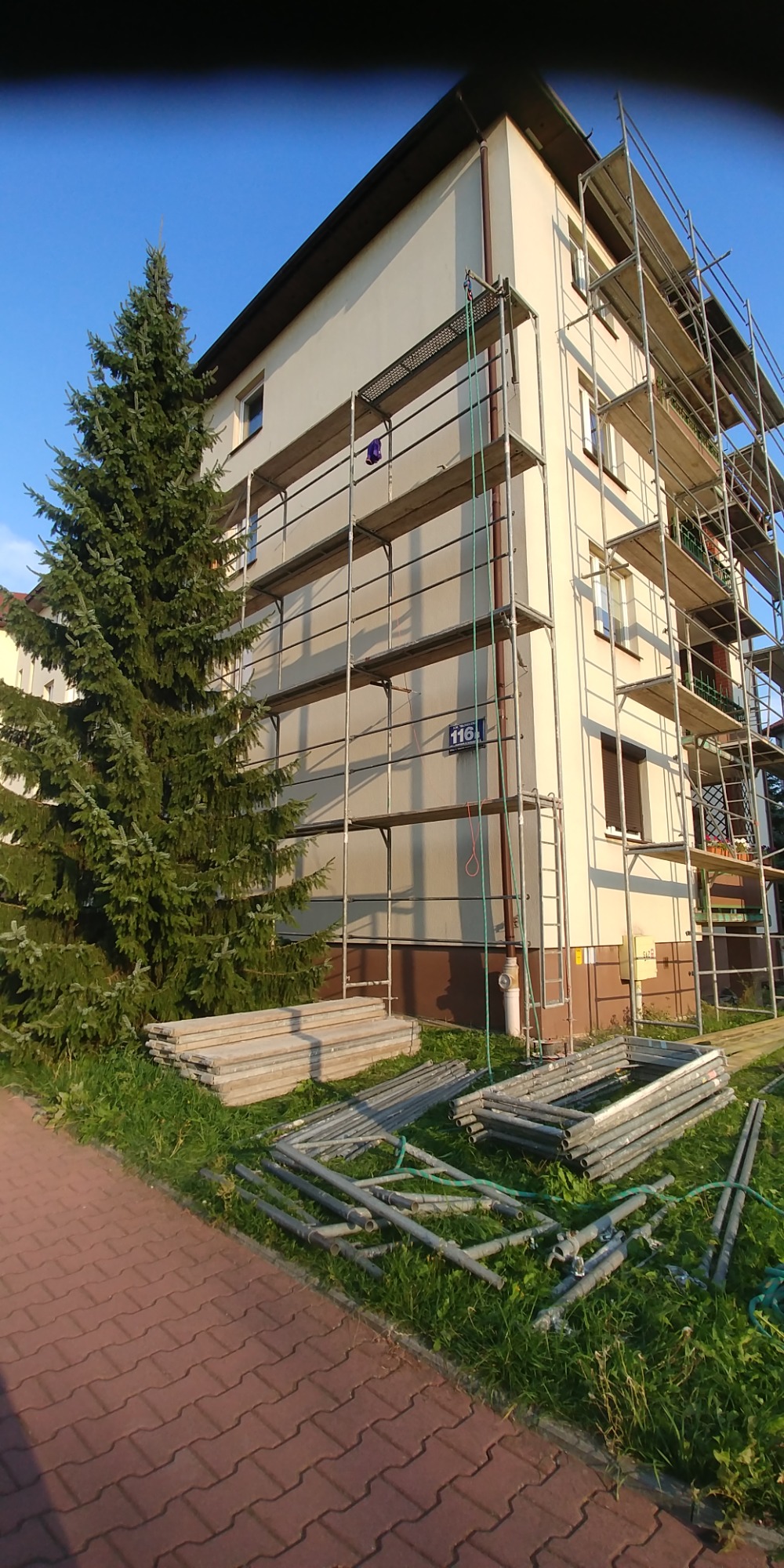 Wielopiętrowy budynek w trakcie prac elewacyjnych, otoczony rusztowaniem, z widocznymi elementami rusztowania składowanymi na trawniku obok ścieżki z kostki brukowej i dużej choinki.