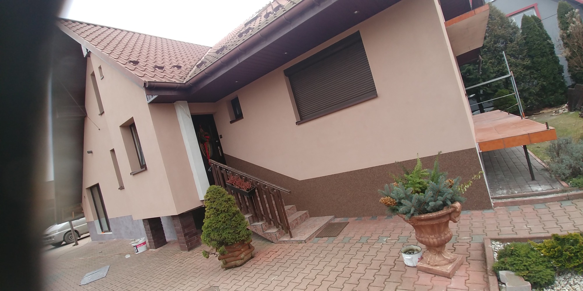 Dwupiętrowy dom z beżową elewacją, brązową stolarką okienną i schodami wejściowymi, otoczony kostką brukową i dekoracyjnymi roślinami w donicach.