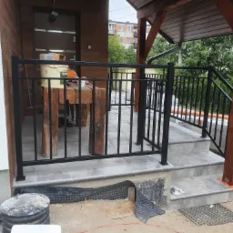 Czarna, metalowa poręcz przy schodach wejściowych do domu w Rzeszowie, widoczne materiały budowlane i niedokończone wykończenie.