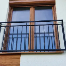 Czarna, metalowa balustrada balkonowa z prostymi prętami, zamontowana na jasnej elewacji budynku z drewnianymi elementami wykończeniowymi okna i dolnej części ściany.
