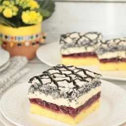 Kwadratowy kawałek ciasta z makiem, kremem i wiśniami, polany czekoladą, na białym talerzu z tłoczeniami, w tle rozmyte dwa inne kawałki i bukiet żółtych kwiatów w ozdobnej doniczce.