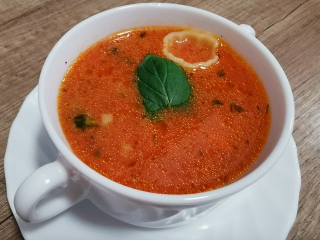 Porcja zupy pomidorowej w białej filiżance, udekorowana listkiem bazylii i małym makaronem, ustawiona na drewnianym stole.