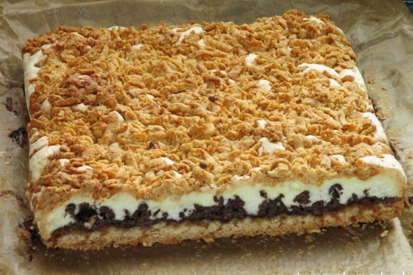 Kawałek domowego ciasta z kruszonką, kremem i czekoladowym spodem, ułożony na papierze do pieczenia.