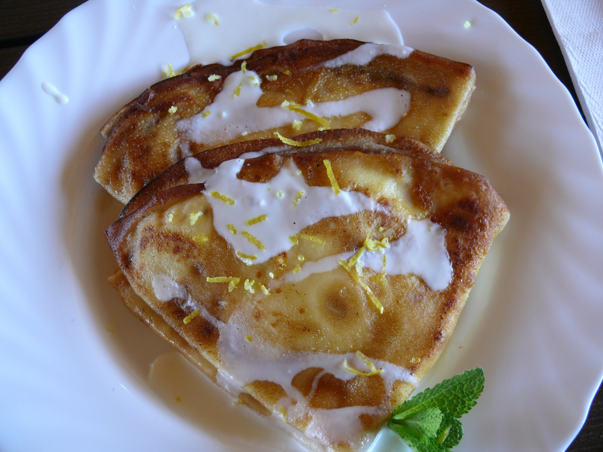 Dwa złożone naleśniki polane białym sosem i posypane skórką cytrynową, udekorowane gałązką mięty na białym talerzu.