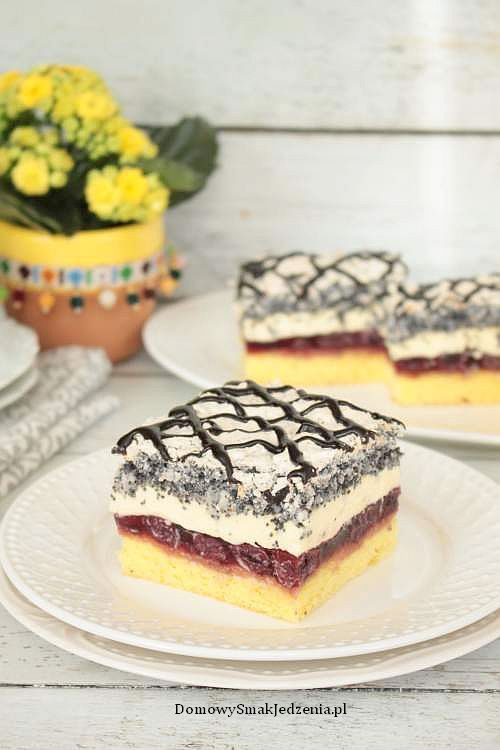 Kwadratowy kawałek ciasta z makiem, kremem i wiśniami, polany czekoladą, na białym talerzu z tłoczeniami, w tle rozmyte dwa inne kawałki i bukiet żółtych kwiatów w ozdobnej doniczce.