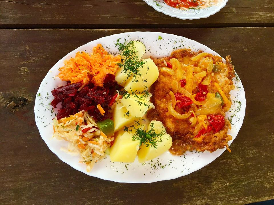 Tradycyjny obiad na białym talerzu: kotlet schabowy z duszoną papryką i cebulą, ziemniaki posypane koperkiem, surówka z marchwi, buraczków i kapusty, widoczne na tle drewnianego stołu.
