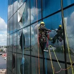 Trzech alpinistów przemysłowych myje okna na wysokościowcu z widokiem na miasto i port, odbicie chmur w szklanej fasadzie.